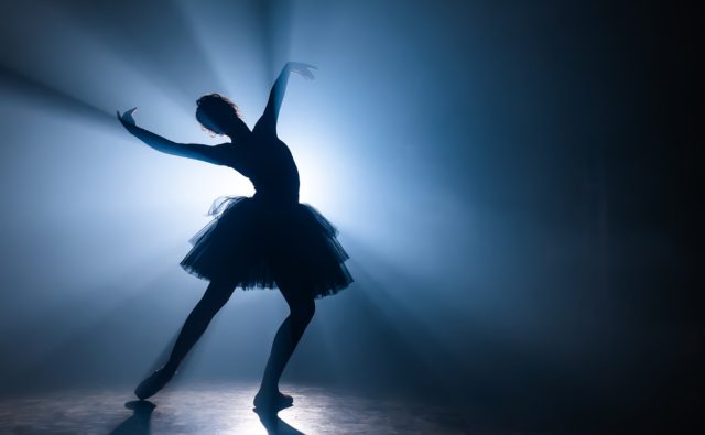 Solo performance by ballerina in tutu dress against backdrop of luminous neon spotlight in theater.